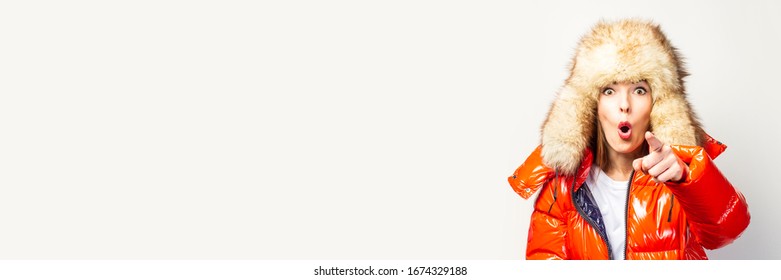 Happy Young Girl In A Red Jacket And A Fur Hat, Looks At The Camera And Points A Finger In Front Of Him On A Light Background. Concept Gestures, Advertising, Fashion. Baner