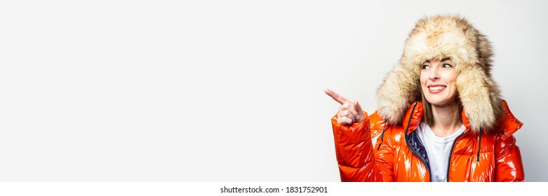 Happy Young Girl In A Red Jacket And A Fur Hat, Looks Away And Points A Finger At A Light Background. Concept Gestures, Advertising, Fashion. Baner