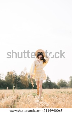 Happy young girl running through a yellow field on a summer day. girl in yellow dress and straw hat with a bouquet of wheat. sheared golden wheat spikelets. Agricultural texture