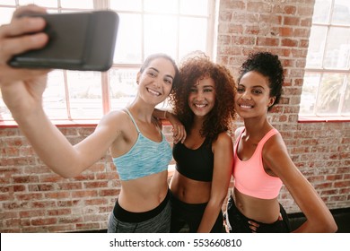 Happy Young Girls In Sportswear Taking Selfie In Gym. Three Female Friends In Healthclub Taking Selfie With Mobile Phone.