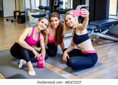 Happy Young Girls In Sportswear Taking Selfie In Gym. Three Female Friends In Healthclub Taking Selfie With Mobile Phone.