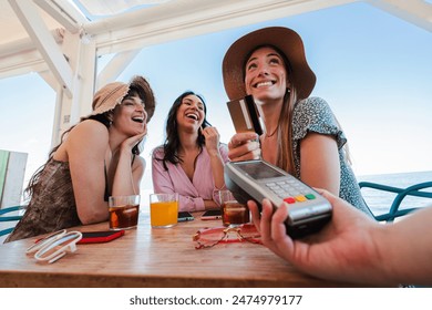 Happy young group of only women paying the bill with a contactless credit card in cocktail beach bar. Female smiling holding a creditcard and giving a payment transaction to the cashier on vacations - Powered by Shutterstock