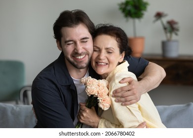 Happy young grown up 30s son cuddling middle aged retired mother, congratulating with birthday or women's day at home, presenting flowers. Bonding two generations family celebrating special occasion.