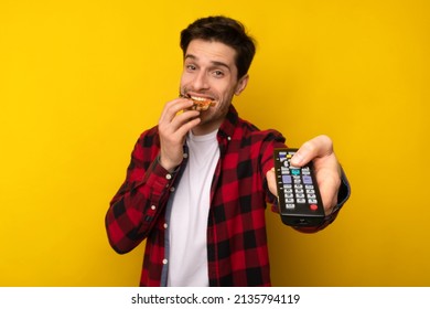 Happy Young Guy Eating Pizza Watching TV Switching Channels With Television Remote Control Posing On Yellow Studio Background, Looking At Camera, Selective Focus On Hand. Junk Food Overeating Concept