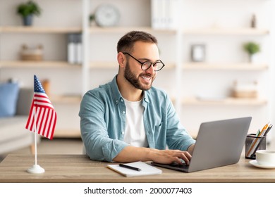 Happy Young Guy With Flag Of The US Working At Desk With Laptop In Home Office. Millennial Male Student Learning English Online For Foreign Job, Education Or Emigration To America