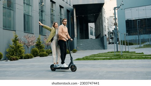 Happy young guy and girl ride on electric scooters near business center at street outdoors, after work. Lifestile concept.