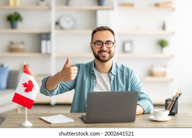 Happy young guy sitting at table with flag of Canada, using laptop computer, showing thumb up gesture at home. Modern online foreign education, emigration and citizenship, working abroad concept