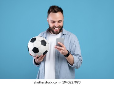 Happy young guy with soccer ball using smartphone, winning sports bet, rooting for his favorite team on blue background