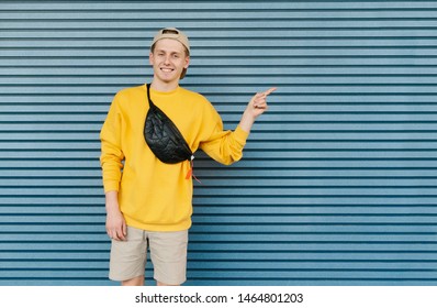 Happy Young Guy In Stylish Clothes, Caps And Fanny Pack Stands On The Background Of A Blue Wall And Shows His Finger On A Blank Space. Amazed Young Man Shows A Hand On Copy Space