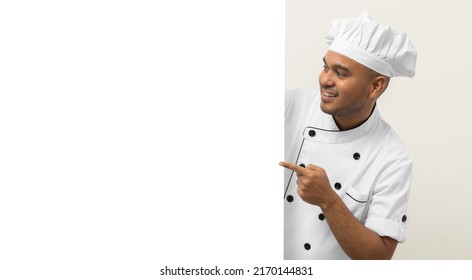 Happy Young Handsome Asian Man Chef In Uniform Looks Out From Behind An Empty Whiteboard. Cooking Indian Man Hiding Behind Big Blank Billboard For Advertising Text Menu In Kitchen And Restaurant.