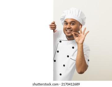 Happy Young Handsome Asian Man Chef In Uniform Looks Out From Behind An Empty Whiteboard. Cooking Indian Man Hiding Behind Big Blank Billboard For Advertising Text Menu In Kitchen And Restaurant.