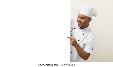 Happy Young Handsome Asian Man Chef In Uniform Looks Out From Behind An Empty Whiteboard. Cooking Indian Man Hiding Behind Big Blank Billboard For Advertising Text Menu In Kitchen And Restaurant.