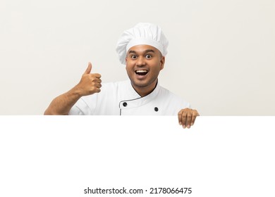 Happy Young Handsome Asian Man Chef In Uniform Looks Out From Behind An Empty Whiteboard. Cooking Indian Man Hiding Behind Big Blank Billboard For Advertising Text Menu In Kitchen And Restaurant.