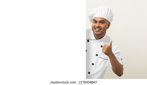 Happy Young Handsome Asian Man Chef In Uniform Looks Out From Behind An Empty Whiteboard. Cooking Indian Man Hiding Behind Big Blank Billboard For Advertising Text Menu In Kitchen And Restaurant.