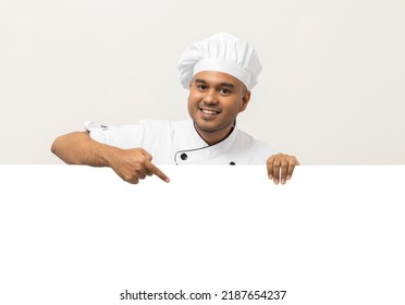 Happy Young handsome asian man chef in uniform looks out from behind an empty whiteboard. Cooking indian man hiding behind big blank billboard for advertising text menu in kitchen and restaurant.