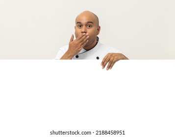 Happy Young Handsome Asian Man Chef In Uniform Looks Out From Behind An Empty Whiteboard. Cooking Indian Man Hiding Behind Big Blank Billboard For Advertising Text Menu In Kitchen And Restaurant.