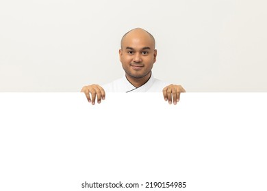 Happy Young Handsome Asian Man Chef In Uniform Looks Out From Behind An Empty Whiteboard. Cooking Indian Man Hiding Behind Big Blank Billboard For Advertising Text Menu In Kitchen And Restaurant.