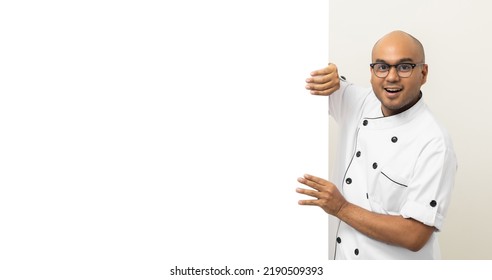 Happy Young Handsome Asian Man Chef In Uniform Looks Out From Behind An Empty Whiteboard. Cooking Indian Man Hiding Behind Big Blank Billboard For Advertising Text Menu In Kitchen And Restaurant.