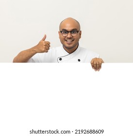 Happy Young Handsome Asian Man Chef In Uniform Looks Out From Behind An Empty Whiteboard. Cooking Indian Man Hiding Behind Big Blank Billboard For Advertising Text Menu In Kitchen And Restaurant.