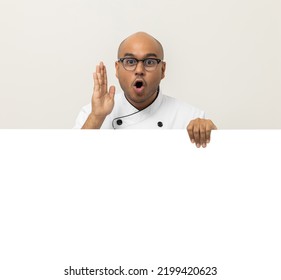 Happy Young Handsome Asian Man Chef In Uniform Looks Out From Behind An Empty Whiteboard. Cooking Indian Man Hiding Behind Big Blank Billboard For Advertising Text Menu In Kitchen And Restaurant.