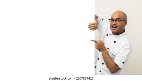 Happy Young Handsome Asian Man Chef In Uniform Looks Out From Behind An Empty Whiteboard. Cooking Indian Man Hiding Behind Big Blank Billboard For Advertising Text Menu In Kitchen And Restaurant.