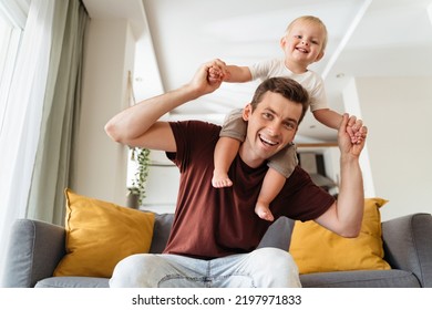 Happy Young Handsome Dad Carrying His Baby Boy On Shoulders Sitting On Sofa Posing In Front Of Camera In Living-room, Both Smiling And Having Fun While Mom Resting. Father Having Fun With His Toddler