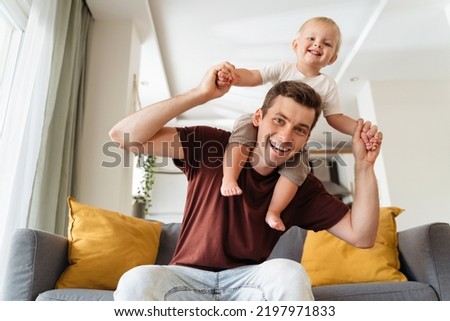 Happy young handsome dad carrying his baby boy on shoulders sitting on sofa posing in front of camera in living-room, both smiling and having fun while mom resting. Father having fun with his toddler