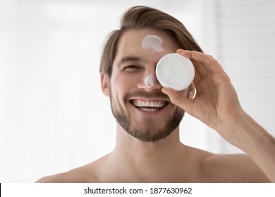 Happy Young Handsome Man Holding Cream, Applying Moisturizing Cosmetics On Face. Smiling Male Blogger Advertising Nourishing Balm, Feeling Satisfied With Anti-wrinkle Effect, Head Shot Close Up.