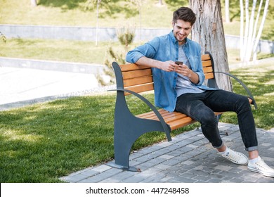 Happy young handsome man sitting on the bench outdoors and using smartphone - Powered by Shutterstock