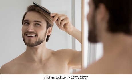 Happy Young Handsome Man Styling Hair With Comb After Morning Shower, Looking In Mirror. Smiling European Guy Getting Ready For Workday Or Preparing For Dating Meeting, Feeling Confident In Bathroom.