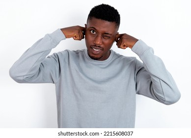 Happy Young Handsome Man Wearing Grey Sweater Over White Background  Ignores Loud Music And Plugs Ears With Fingers Asks To Turn Off Sound