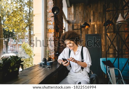 Happy young hipster gen z. Cute woman with afro hair holding smart phone device, using mobile app sitting at outdoor table in modern city cafe. Horizontal view.