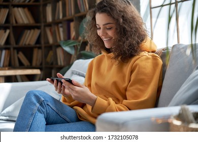 Happy Young Hispanic Latin Teen Girl Sit On Sofa At Home Holding Phone Looking At Screen Watching Social Media Video Content, Movie Or Stream, Video Calling Online In Mobile App Using Smart Phone.