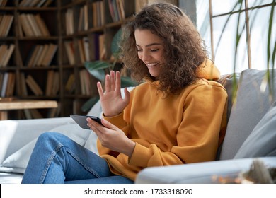 Happy young hispanic latin teen girl sit on sofa at home holding phone looking at screen waving hand video calling distance friend online in mobile chat app using smartphone videochat application.