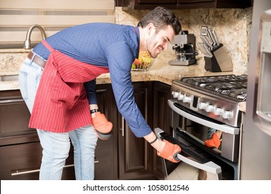 Happy Young Hispanic Man Wearing An Apron And Oven Mitts Opening The Oven Door To Take Out Baked Food