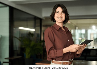 Happy young Hispanic professional business woman using tab standing in office. Happy businesswoman manager female executive leader holding tab at work looking away dreaming of future successful career - Powered by Shutterstock