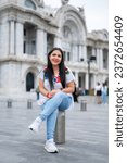Happy young hispanic woman in embroidered top looking at camera while sitting in front of blurred Bellas Fine Arts Palace in Mexico City in daylight enjoying location and wearing traditional clothes