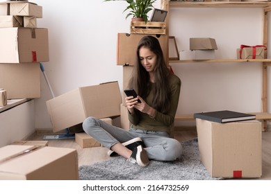 Happy Young Hispanic Woman Using Smartphone In Living Room At New House With Stack Of Cardboard Boxes On Moving Day, Smiling