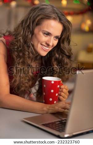 Happy young housewife with cup of hot chocolate using laptop in christmas decorated kitchen