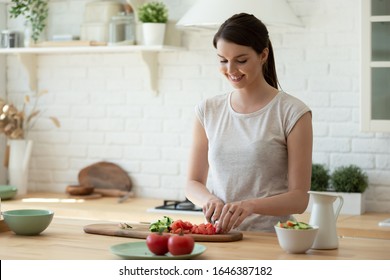 Happy young housewife stand at wooden kitchen counter chop vegetables preparing healthy salad for lunch or dinner, smiling woman cooking in renovated modern home, vegetarian, dieting concept