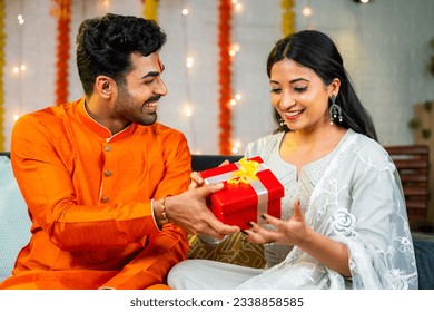 Happy young husband giving present to wife during festival celebration at home - brother giving gift to sister during raksha bandhan.