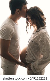 Happy young husband kissing his pregnant wife and holding belly bump. Stylish pregnant family, mom and dad in white pajamas, relaxing at home in light. Fertility concept. True happiness