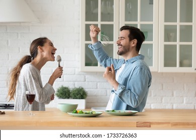 Happy Young Husband And Wife Laugh Sing Using Kitchen Appliances Cooking Together At Home, Overjoyed Smiling Millennial Couple Have Fun Preparing Food Enjoying Family Leisure Weekend Indoors