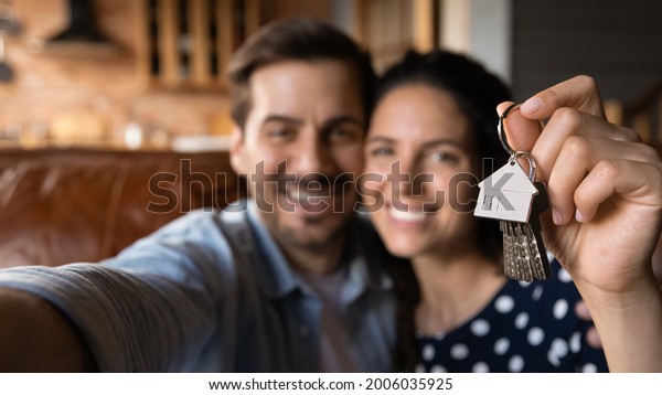 Happy young husband and wife showing key at\
camera, taking selfie. Property buyers, tenants celebrating\
mortgage approval, rent apartment, buying first house. New home,\
real estate concept