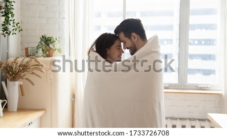 Happy young husband and wife stand hugging cuddling wrapped in cover enjoy tender sweet moment at home, loving millennial couple warm up together in kitchen share spend romantic weekend indoors