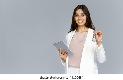 Happy Young Indian Business Woman Professional Realtor Real Estate Agent Wear Suit Looking At Camera Holding Key To New Property House And Digital Tablet Isolated On Grey Studio Background Copy Space