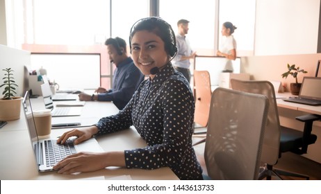 Happy young indian business woman call center agent looking at camera posing in customer service support office, smiling female telemarketer operator wear wireless headset work on computer, portrait