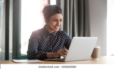Happy young indian business woman entrepreneur using computer looking at screen working in internet sit at office desk, smiling hindu female professional employee typing email on laptop at workplace