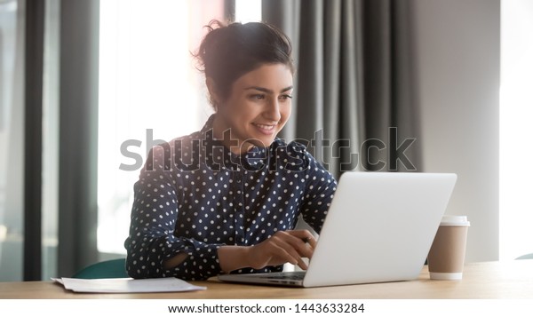 Happy young indian business woman entrepreneur\
using computer looking at screen working in internet sit at office\
desk, smiling hindu female professional employee typing email on\
laptop at workplace