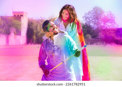 Happy young Indian couple having fun playing with colorful powder color or gulal celebrating holi festival at park outdoor.
